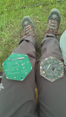 Photo of two hexagonal PCBs lying on a person&rsquo;s knees. The right PCB is covered in components